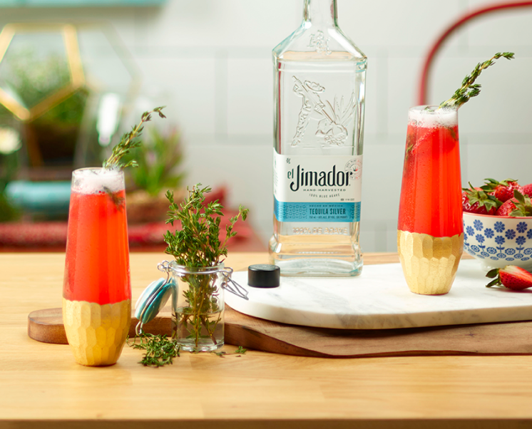 el Jimador Strawberry Bellini cocktails on a table with rosemary herbs, strawberry slices and el Jimador Silver bottle.