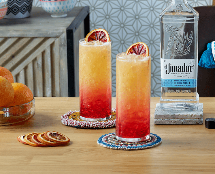 el Jimador Tequila Sunrise cocktails on a table with a burnt orange slices and el Jimador Silver bottle.