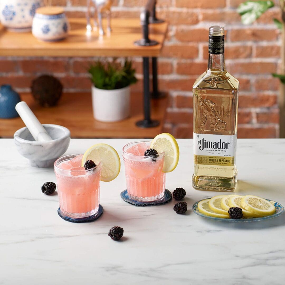 An image of two cocktails on a marble counter with a bottle of el Jimador Reposado tequila, lemon slices, blackberries, and a mortar and pestle