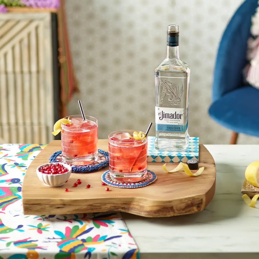 An image of two cocktails on a butcher block along with a bottle of el Jimador Silver tequila and a small bowl of pomegranate