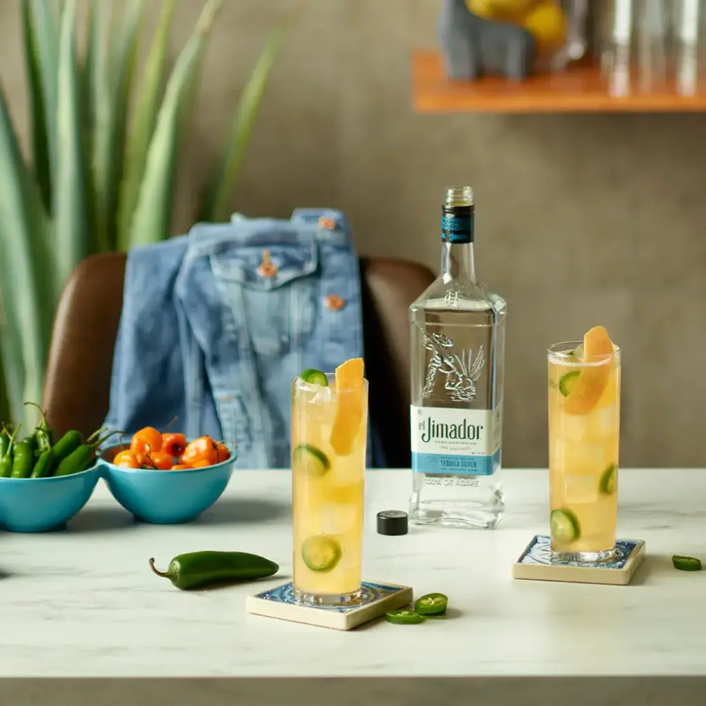 An image of two cocktails on a counter with a bottle of el Jimador Silver tequila and a bowl of green peppers and a bowl of orange peppers