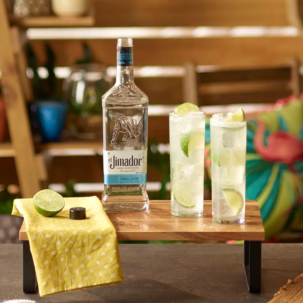 An image of two cocktails on a wooden tray with a bottle of el Jimador Silver tequila and a small yellow napkin with a sliced lime
