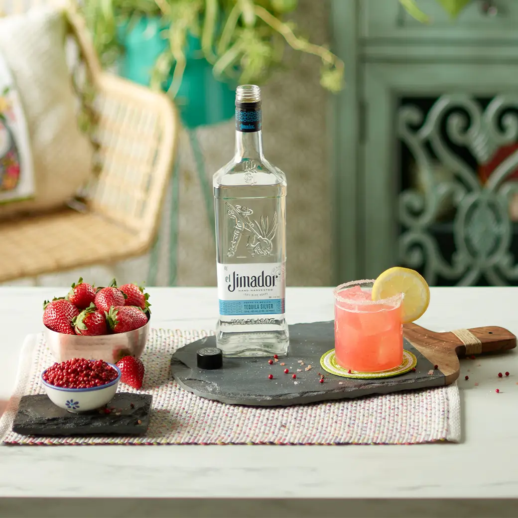 An image of a cocktail on a tray with a bottle of el Jimador Silver tequila along with a bowl of fresh strawberries and a bowl of pomegranate