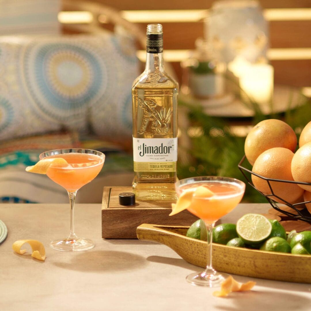 An image of 2 Siesta cocktails on a counter with limes, oranges and a bottle of el Jimador Reposado tequila