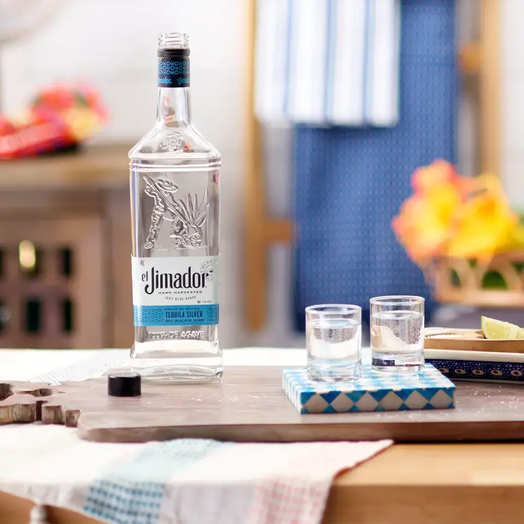 An image of two filled shot glasses on a counter with a lime wedge and a bottle of el Jimador Silver tequila