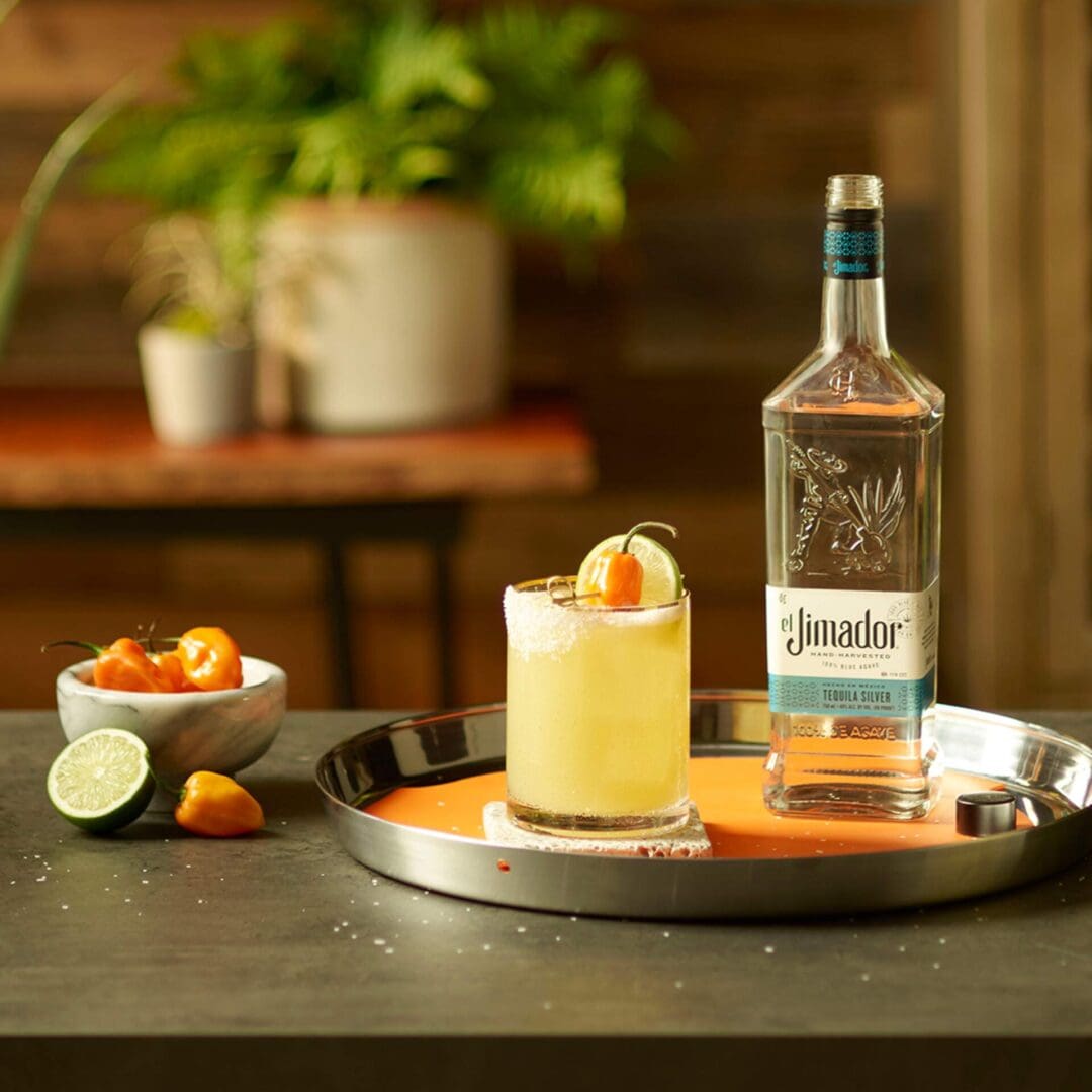An image of 2 spicy margarita cocktails on a counter with habanero peppers, limes and a bottle of el Jimador Silver tequila