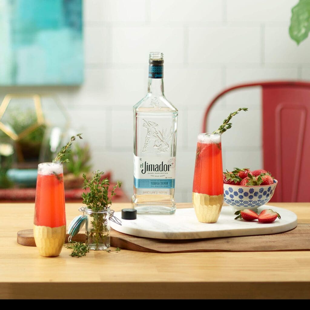 An image of 2 strawberry bellini cocktails on a counter with strawberries, thyme sprigs and a bottle of el Jimador Silver tequila