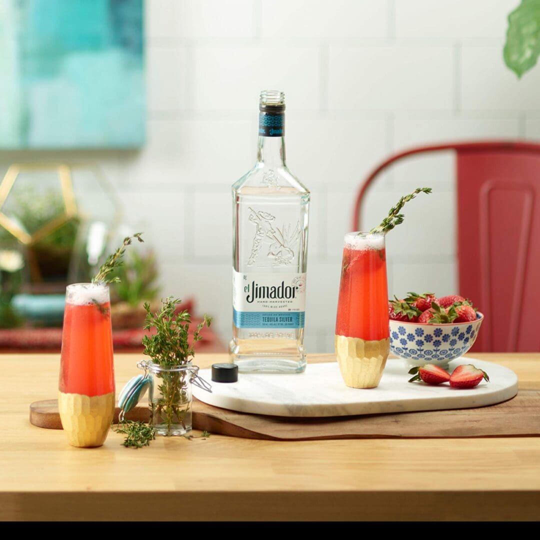 An image of 2 strawberry bellini cocktails on a counter with strawberries, thyme sprigs and a bottle of el Jimador Silver tequila