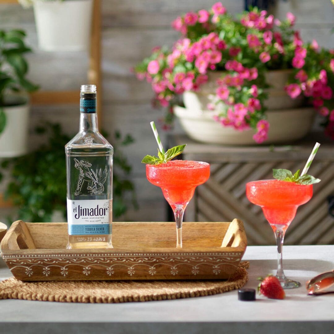 An image of 2 frozen strawberry margarita cocktails on a counter with strawberries, colorful flowers and a bottle of el Jimador Silver tequila