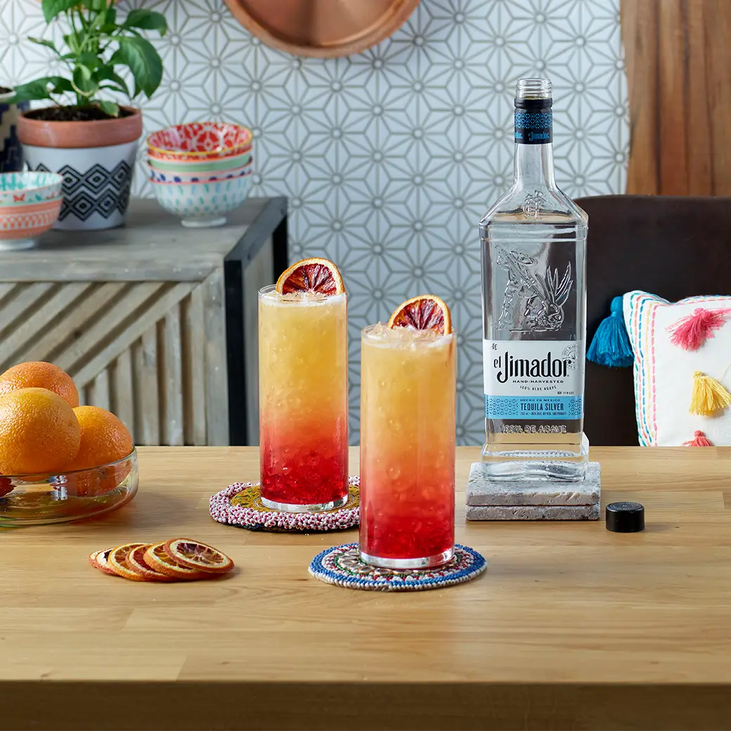 An image of 2 tequila sunrise cocktails on a counter with oranges and a bottle of el Jimador Silver tequila