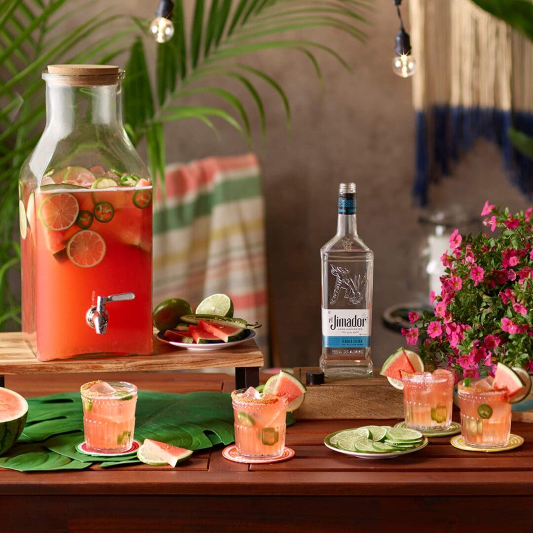 An image of 4 watermelon Picante punch cocktails and a pitcher full of the watermelon picante punch cocktail on a counter with lime wedges, watermelon slices, colorful flowers and a bottle of el Jimador Silver tequila
