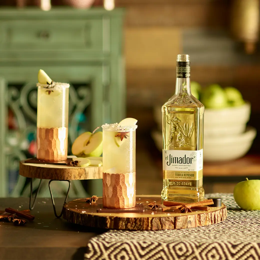 An image of two cocktails on wooden trays with a bottle of el Jimador Reposado tequila and a sliced apple